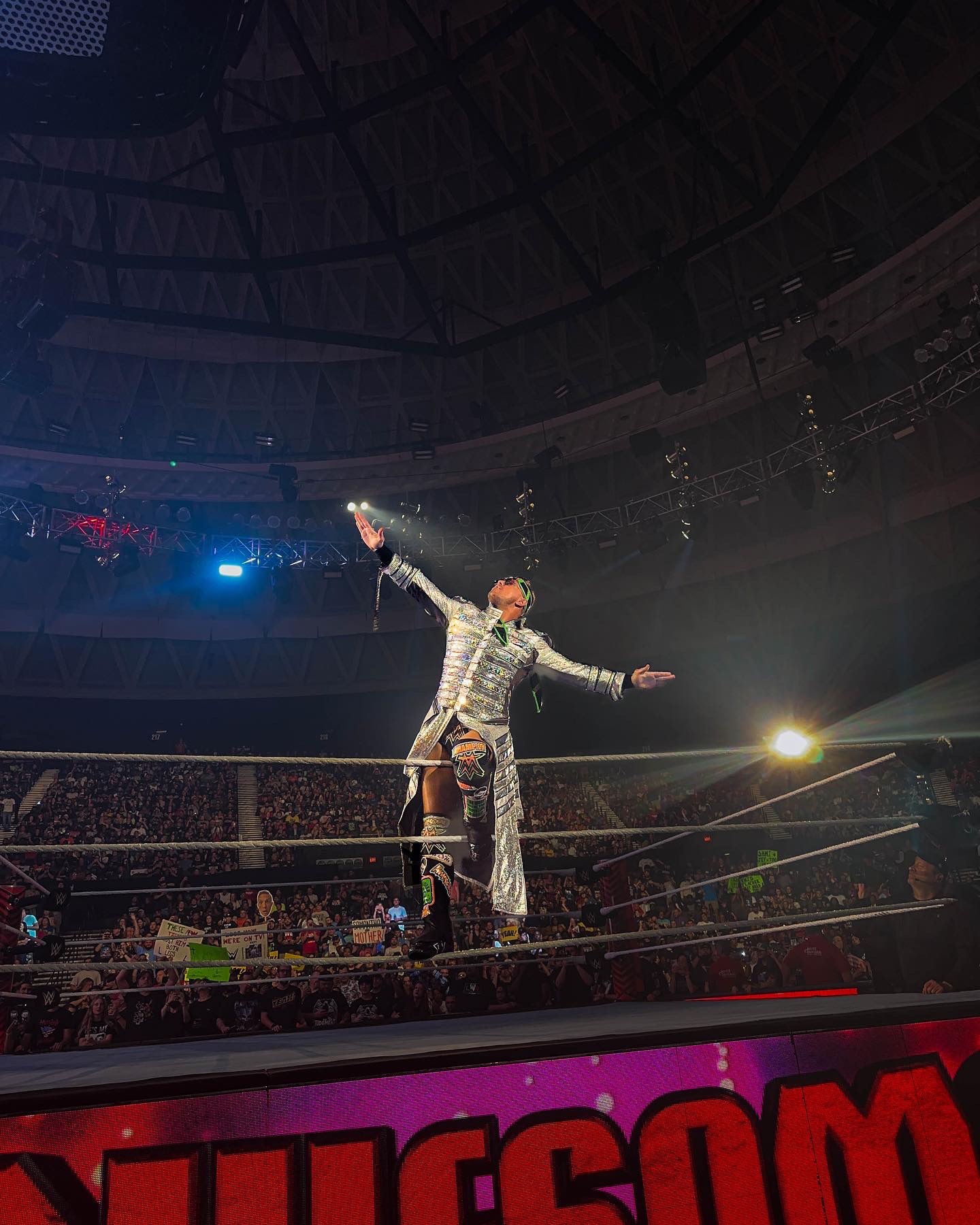 WWE wrestler in a sequined jacket raising his arms on the top rope, championship title on his waist