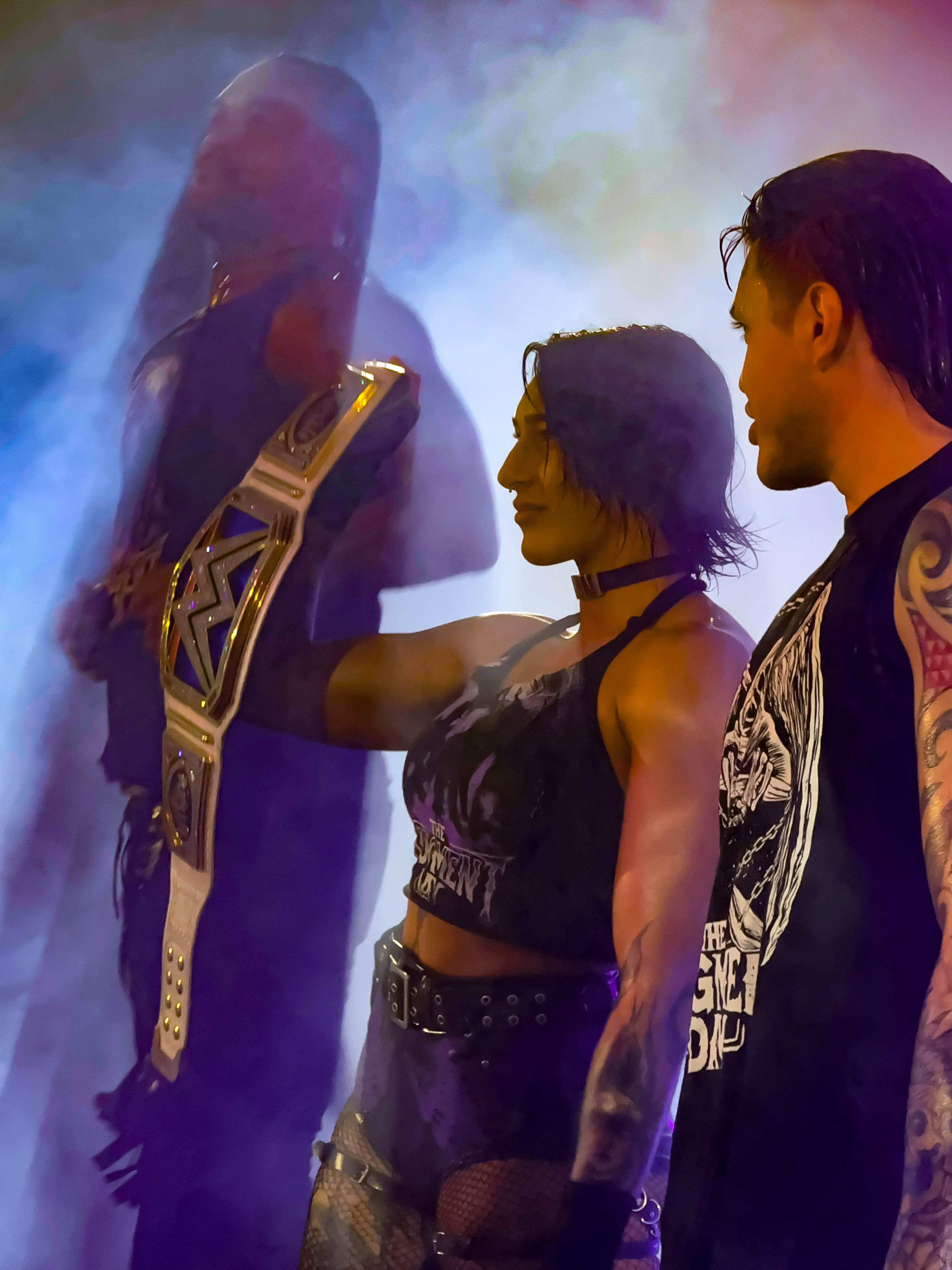 Two WWE wrestlers backstage under colored lighting, one holding a championship title